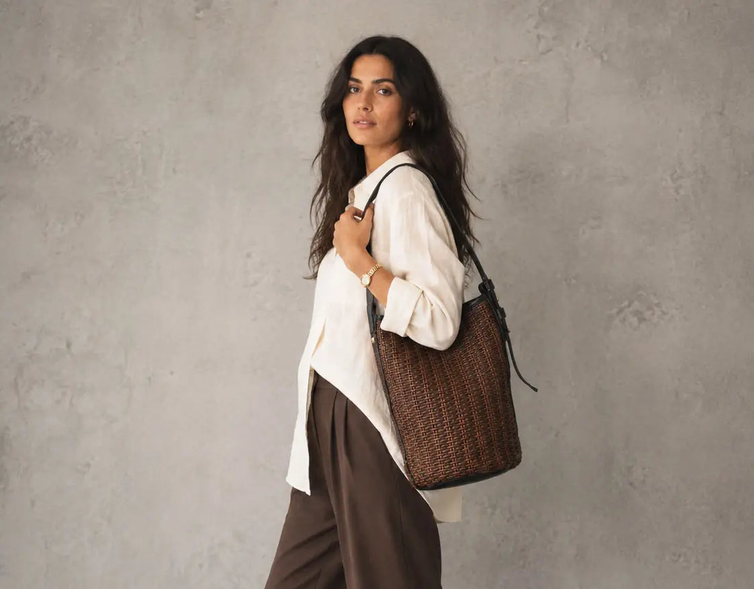A woman wearing a white blouse and brown trousers carries a large brown woven straw tote bag with black trim against a textured gray wall.