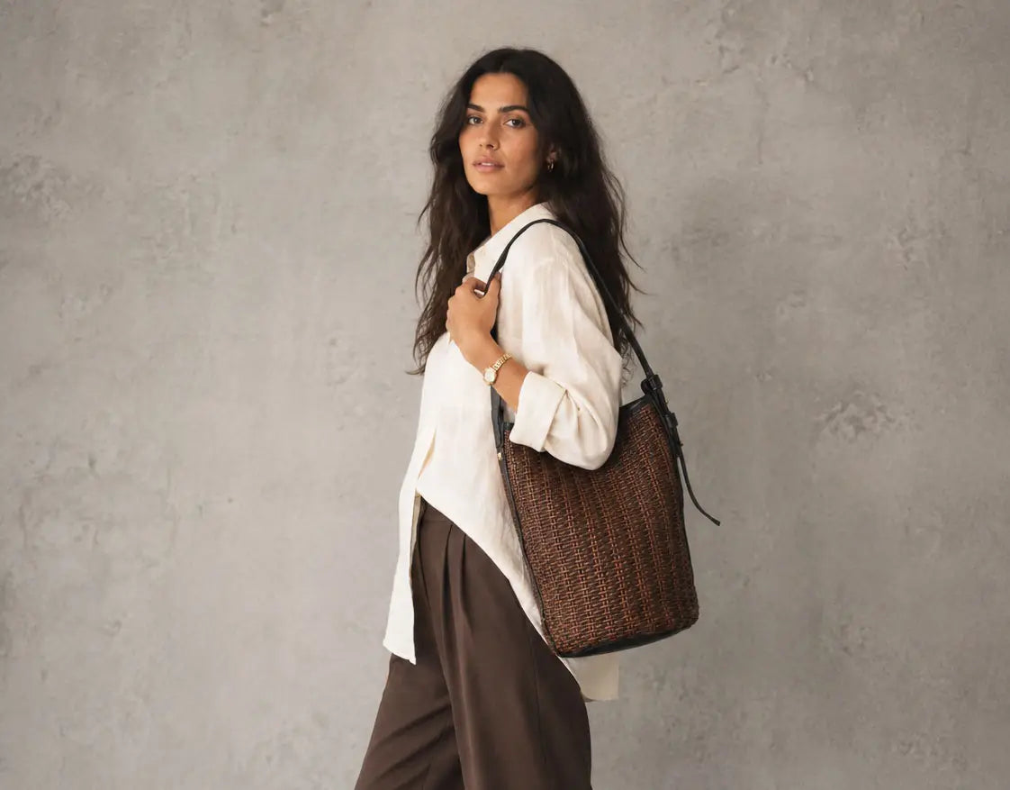 A woman wearing a white blouse and brown trousers carries a large brown woven straw tote bag with black trim against a textured gray wall.