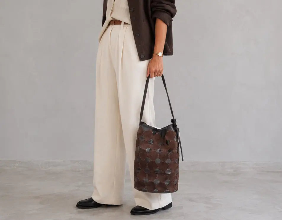 A person wearing cream wide-leg trousers, a dark brown jacket, and black loafers holds a brown patterned bucket bag with black straps against a light gray studio background.