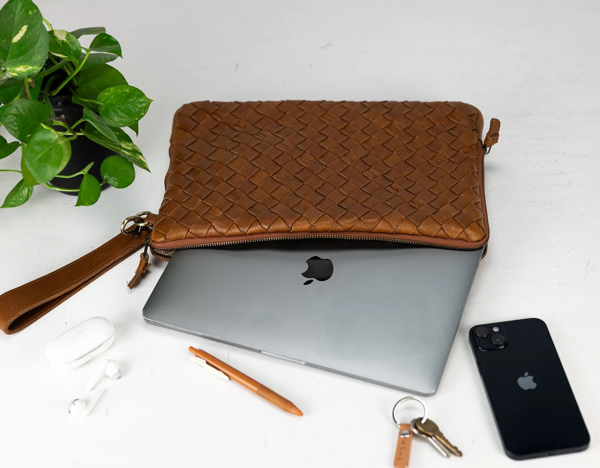 Brown woven clutch with laptop, phone, and accessories on a white surface with a plant.