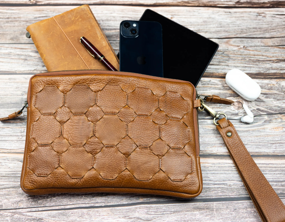 Brown leather clutch with geometric pattern on a wooden surface with phone, pen, and earbuds.