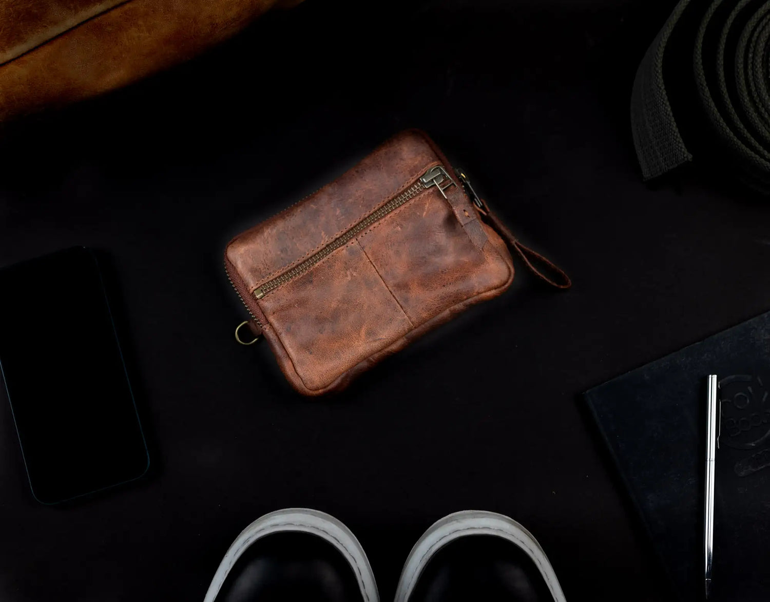 Brown leather pouch on a dark surface with a phone, slippers, and other items.