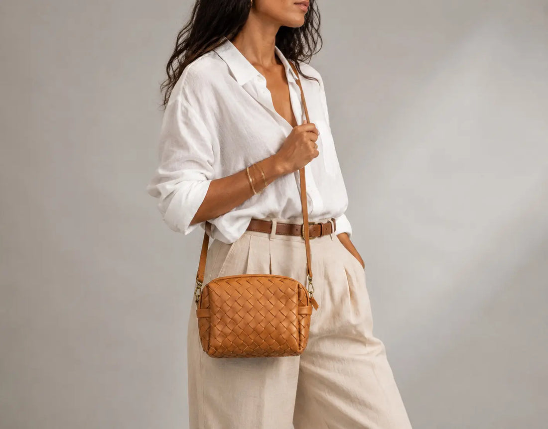 A woman wearing a white button-down shirt and beige trousers carries a small woven tan leather crossbody bag with a brown strap against a neutral gray studio background.