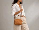 A woman wearing a white button-down shirt and beige trousers carries a small woven tan leather crossbody bag with a brown strap against a neutral gray studio background.