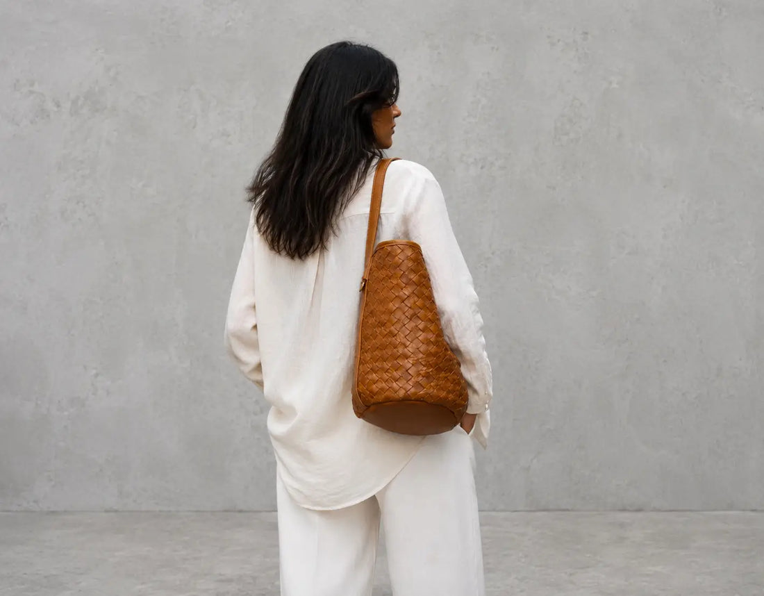 A woman with long black hair is shown from behind wearing a white outfit and carrying a brown woven leather shoulder bag against a textured gray concrete wall.