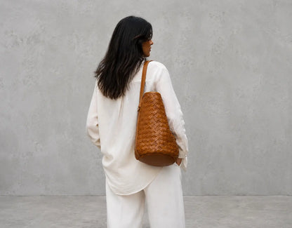 A woman with long black hair is shown from behind wearing a white outfit and carrying a brown woven leather shoulder bag against a textured gray concrete wall.