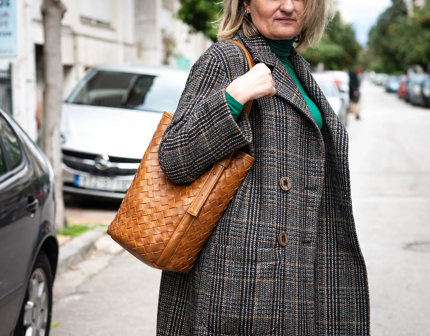 Woman in a plaid coat holding a brown woven bag on a city street.