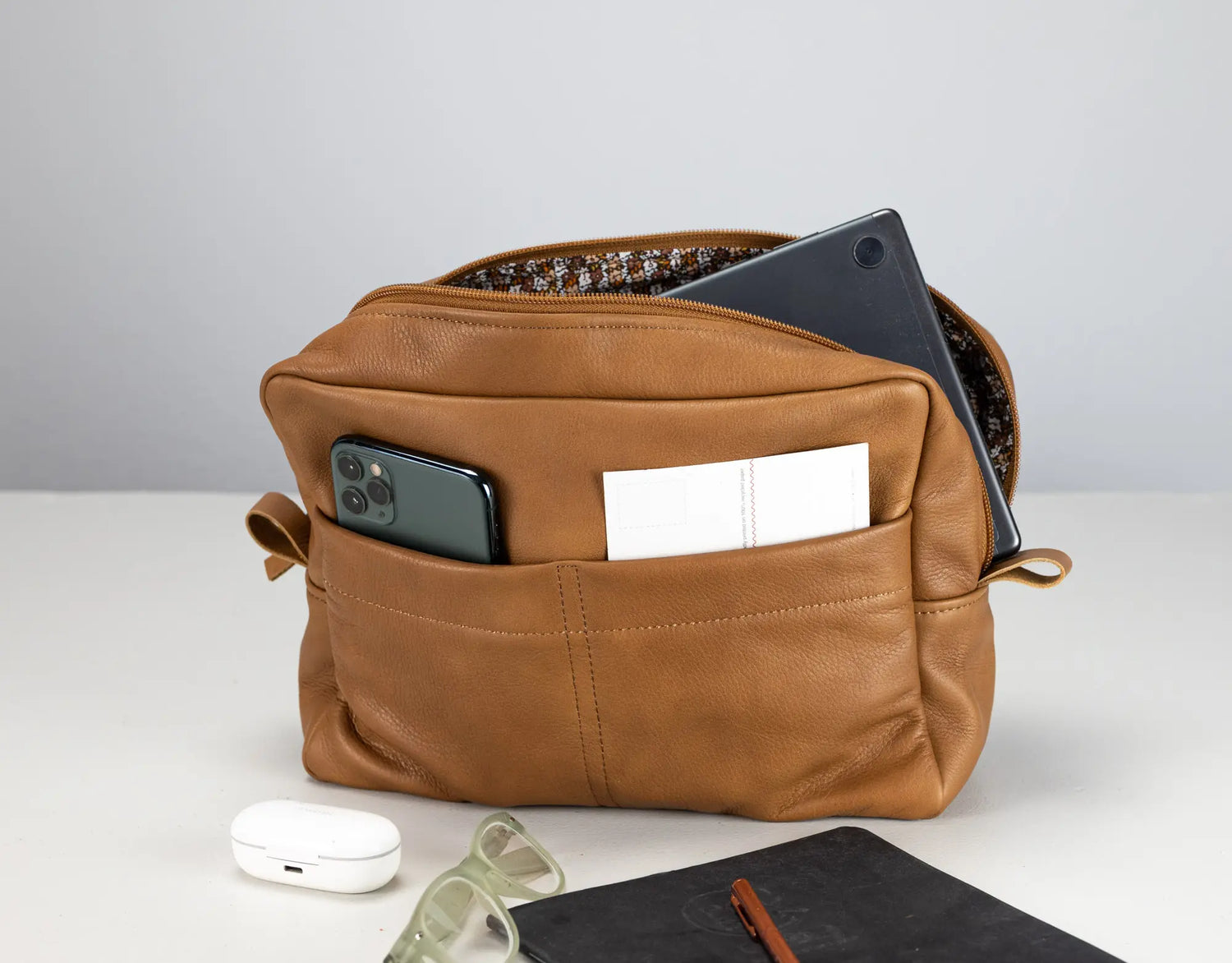 Brown leather bag with phone, notebook, and glasses on a light gray background
