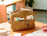 Brown leather bag with compartments on a wooden table, with a hand holding a pen and a cup of coffee.