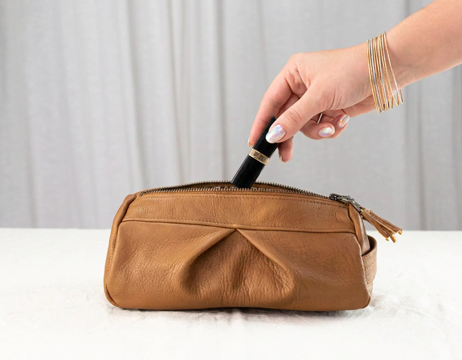 Brown leather clutch with a hand reaching in to place a black item inside, on a light gray background.