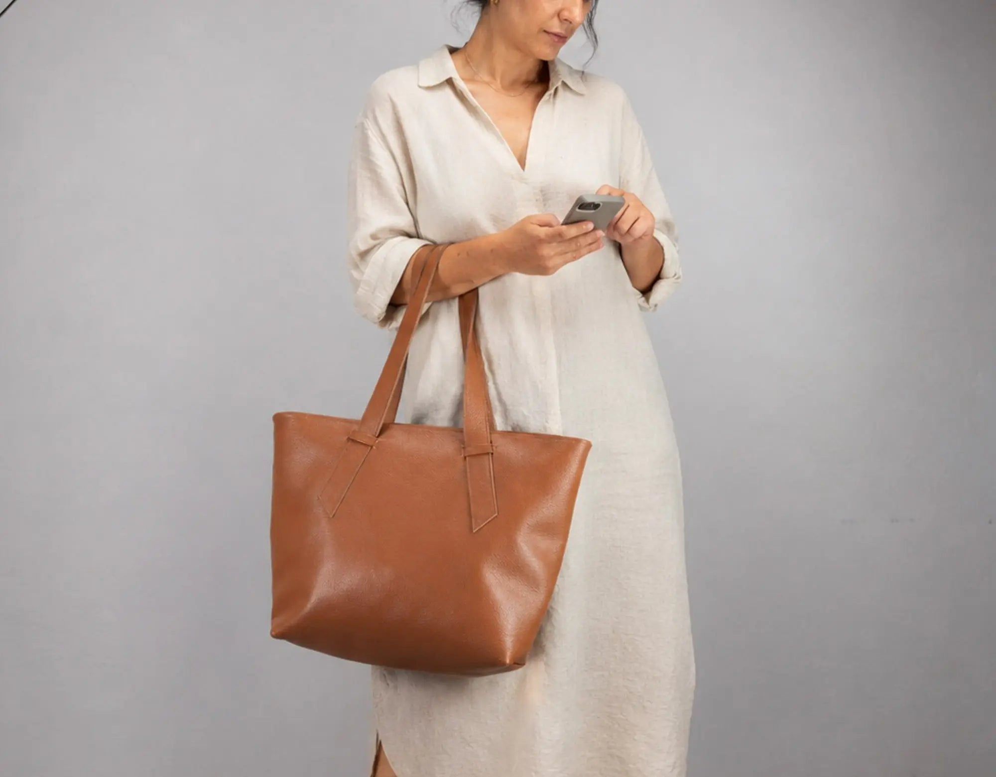 A woman in a cream linen dress holding a brown leather tote bag against a plain gray studio background.