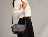 A person wearing a cream sweater and black trousers carries a small black and gray houndstooth crossbody bag with a black leather trim and strap, shown against a neutral gray background.