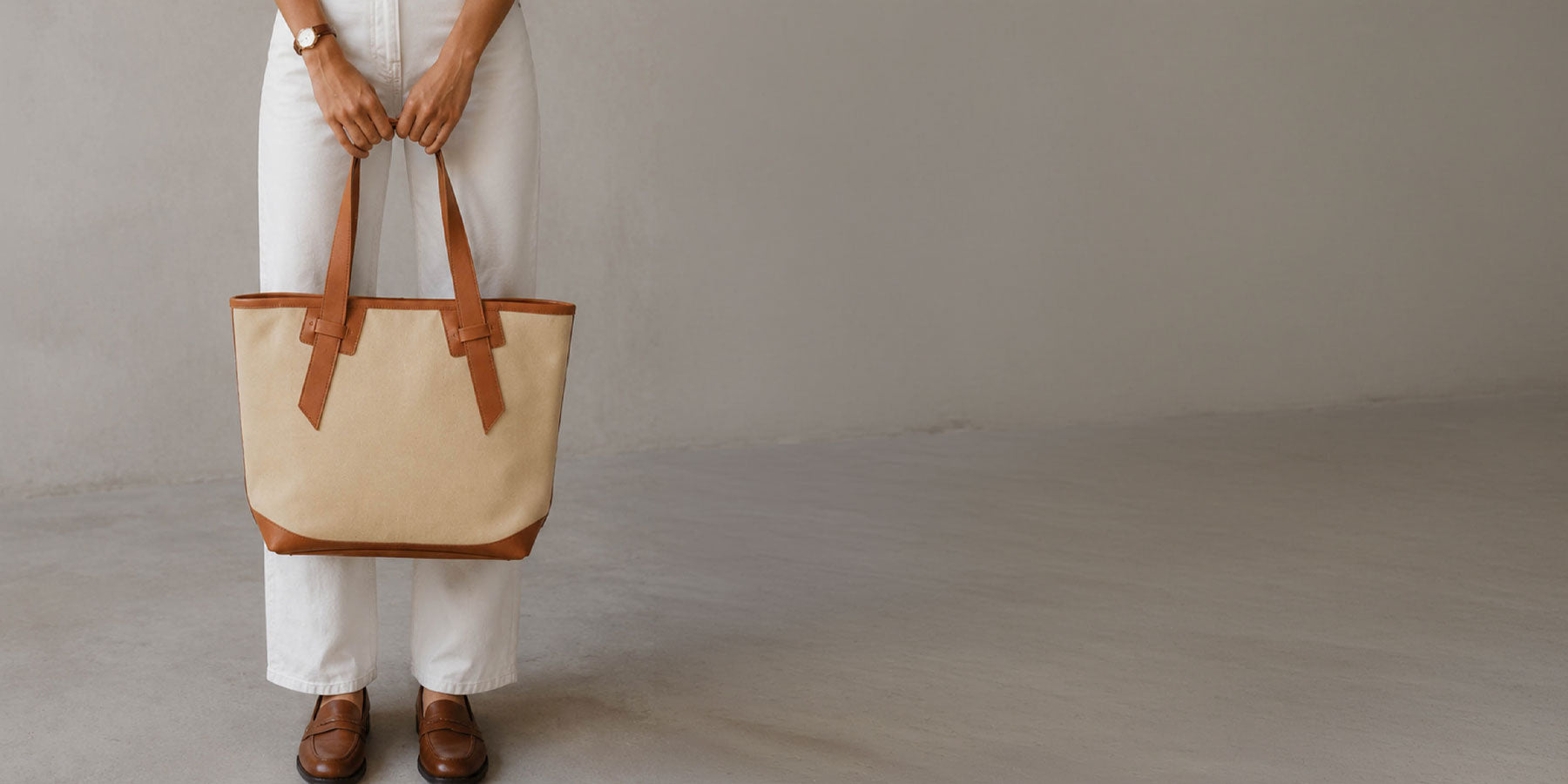 A person wearing white pants and brown shoes holds a large beige canvas tote bag with brown leather trim and handles against a plain light gray studio background.
