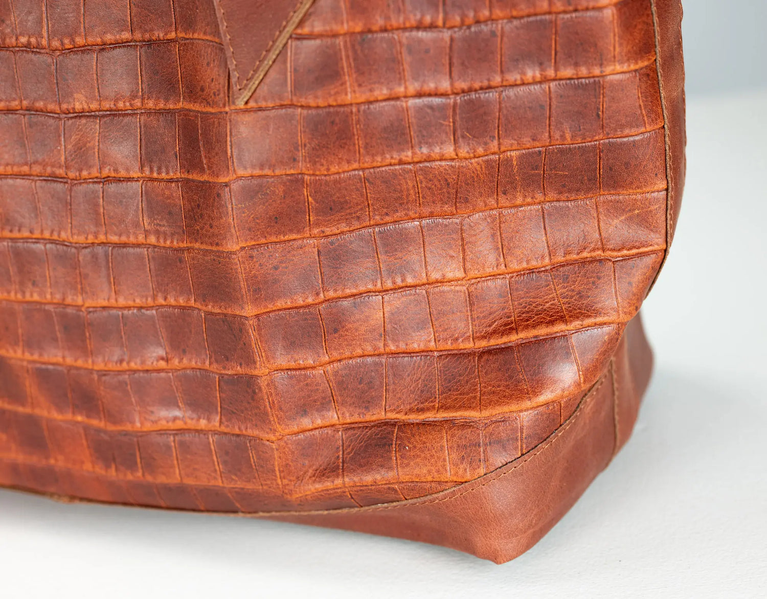 Close-up of a brown crocodile-embossed leather bag corner showing the textured pattern and stitched edge on a light gray background.