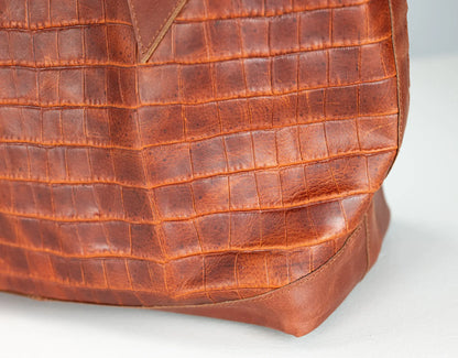Close-up of a brown crocodile-embossed leather bag corner showing the textured pattern and stitched edge on a light gray background.