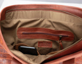Close-up of the interior of a brown leather bag, showing a tan fabric lining, a zippered pocket, a phone in a pocket, and a small &