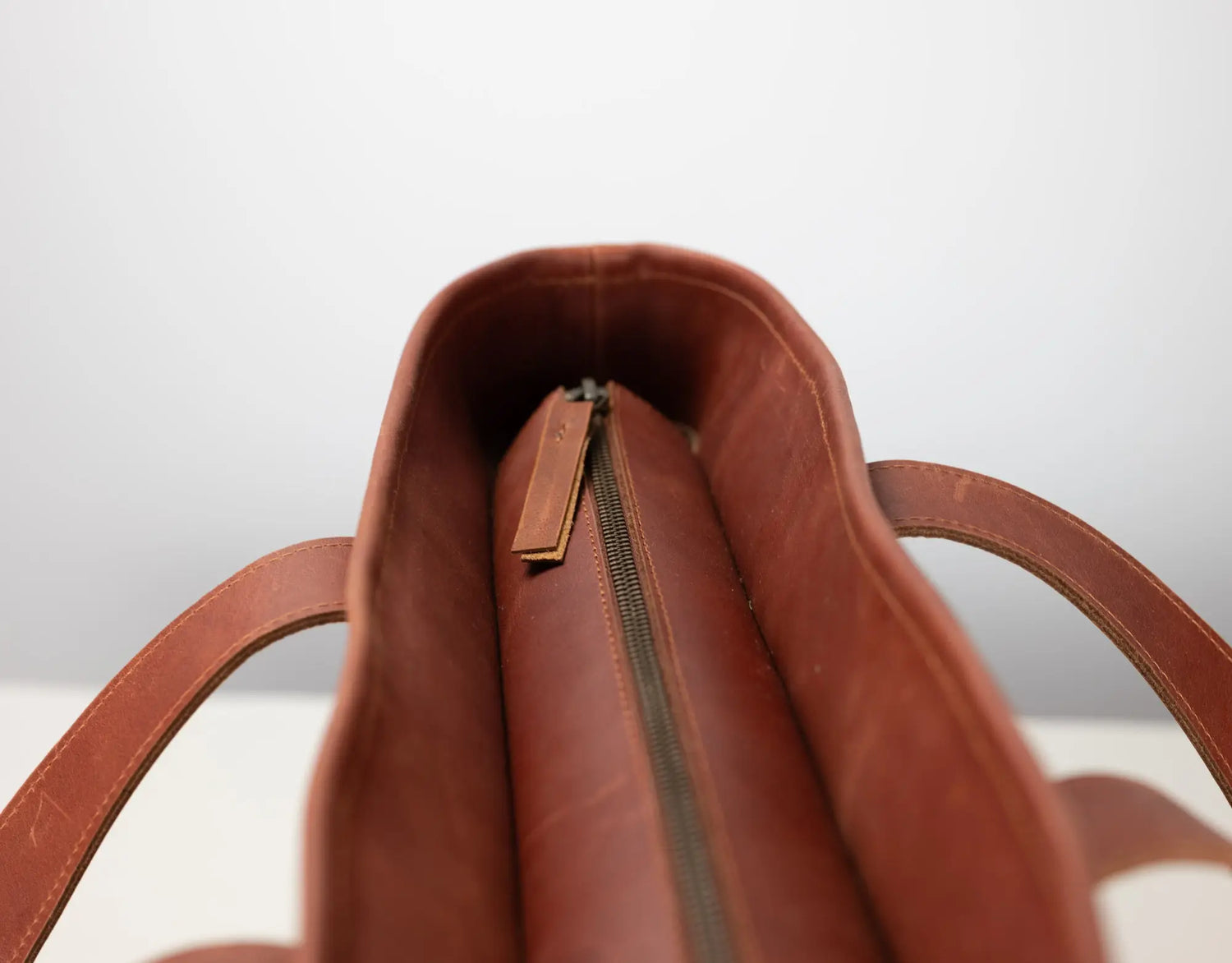 Close-up of the interior top opening of a brown leather bag, showing a zipper closure and part of the strap on a light gray background.