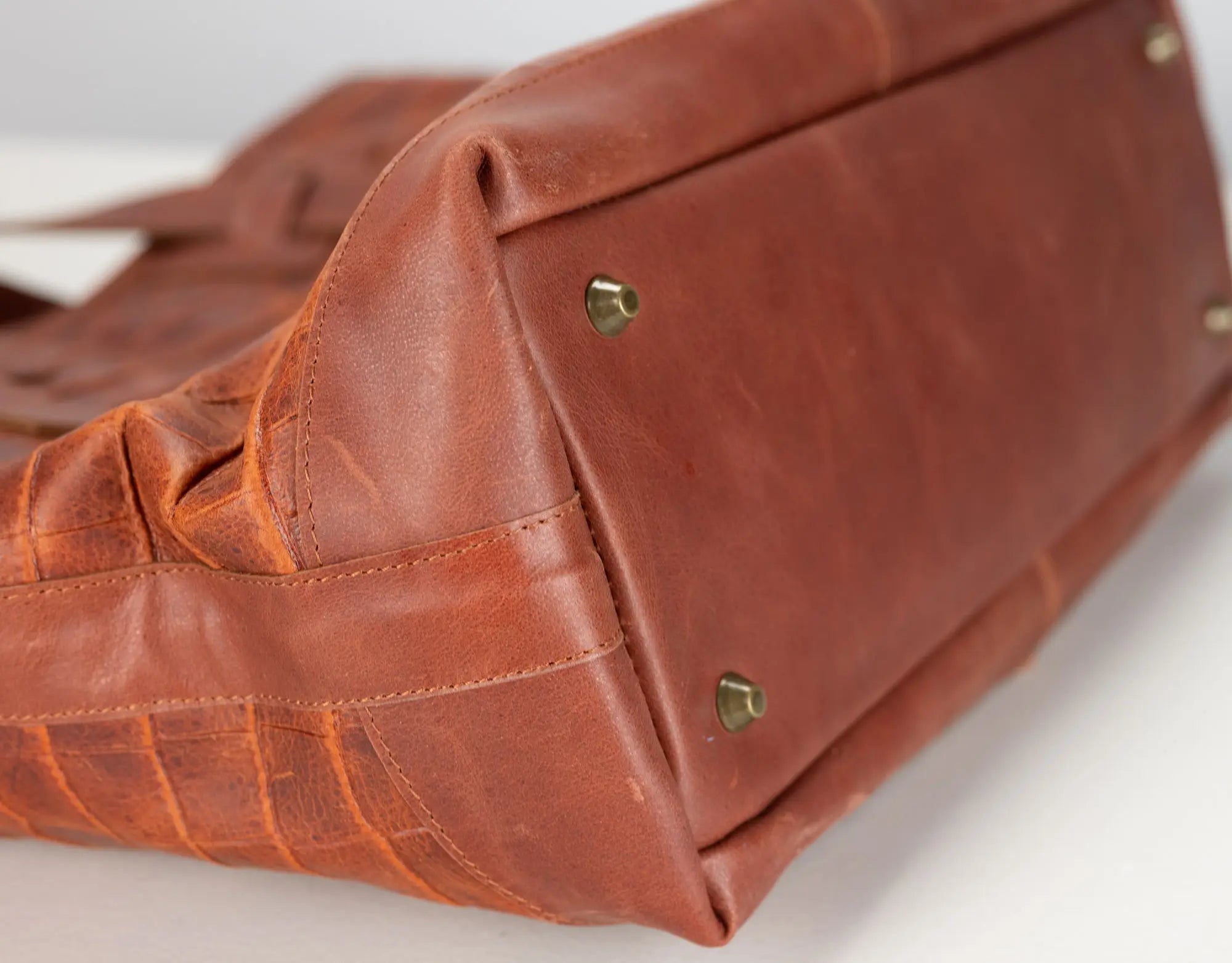 Close-up of a brown leather handbag corner showing stitched seams, a textured side panel, and small brass-tone metal studs on a white surface.