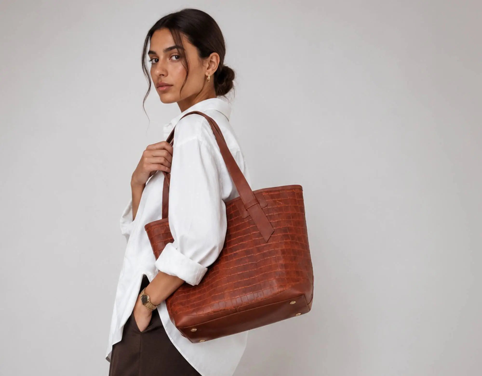 A woman wearing a white button-down shirt and dark brown trousers carries a large brown crocodile-embossed leather tote bag over her shoulder, set against a plain light gray studio background.