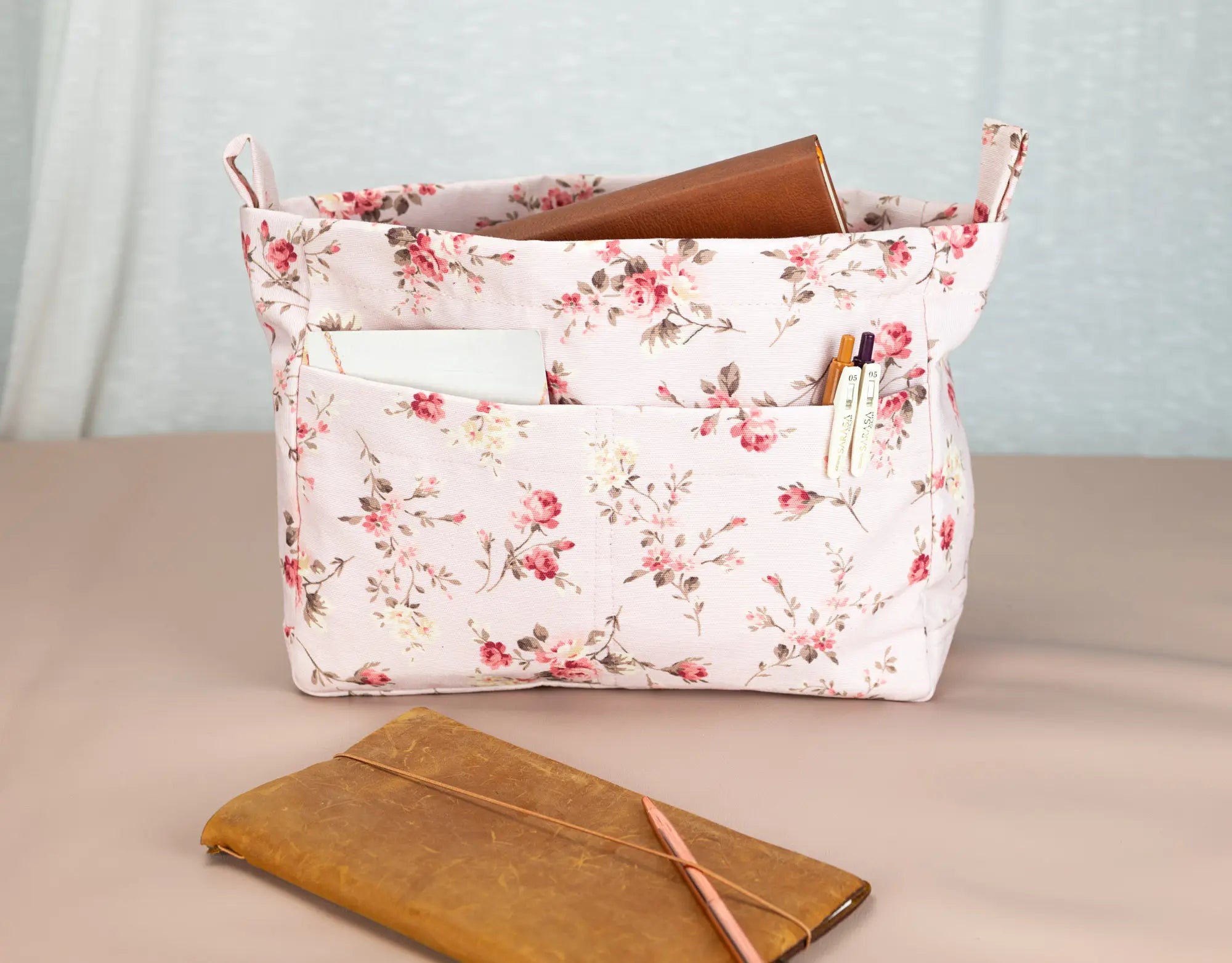 Floral-patterned fabric basket with a brown notebook and pencils on a beige surface.