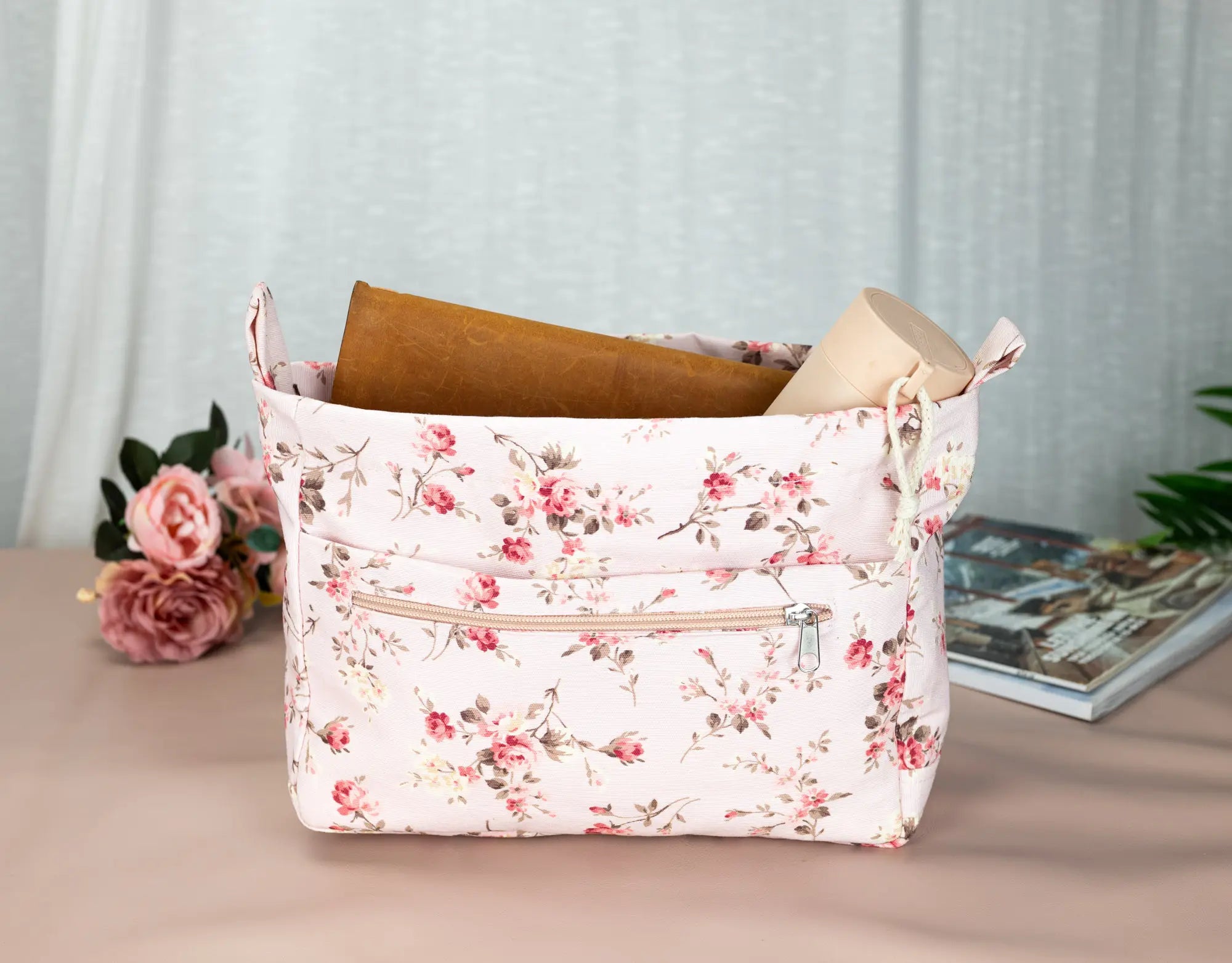 Floral-patterned storage basket with books on a light surface, with flowers and a magazine in the background.