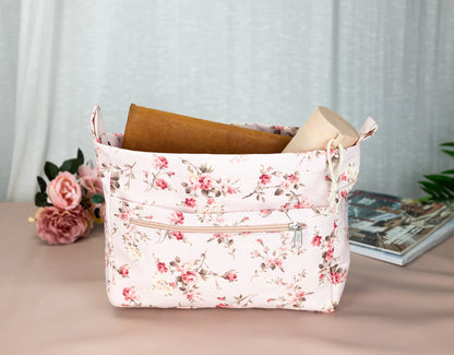 Floral-patterned storage basket with books on a light surface, with flowers and a magazine in the background.