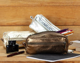 Bronze pouch, silver pencil case, ink bottle, and pen on a wooden surface
