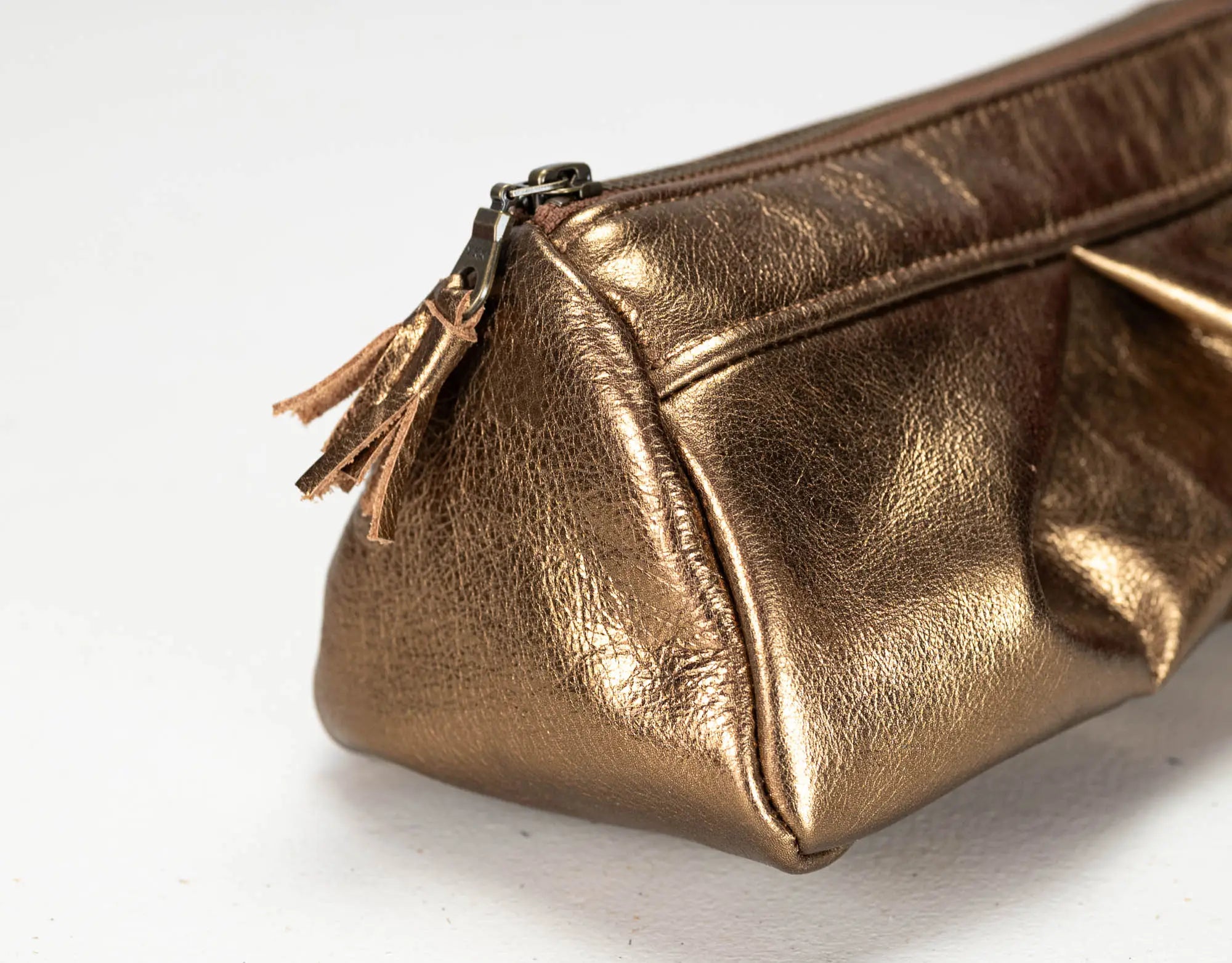 A close-up of a metallic bronze leather cosmetic pouch with a matching leather tassel zipper pull, set against a plain white background.