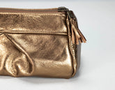 A close-up of a metallic gold leather cosmetic pouch with a top zipper and a matching leather tassel pull, set against a plain light gray background.
