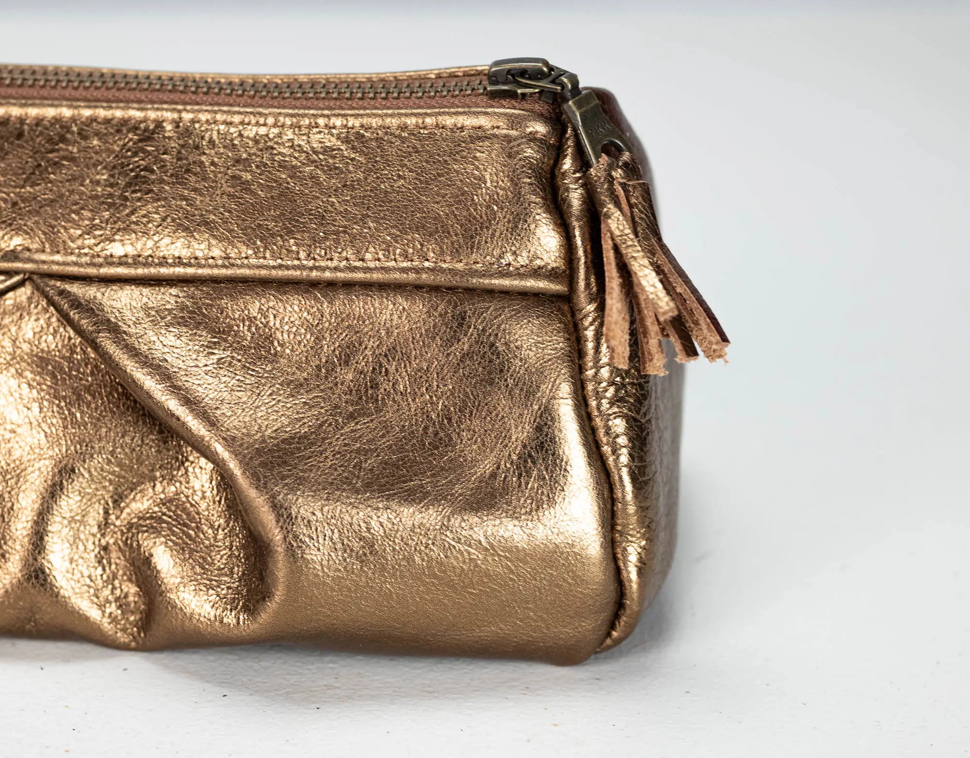 A close-up of a metallic gold leather cosmetic pouch with a top zipper and a matching leather tassel pull, set against a plain light gray background.