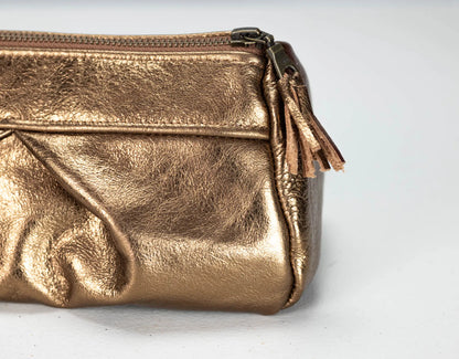 A close-up of a metallic gold leather cosmetic pouch with a top zipper and a matching leather tassel pull, set against a plain light gray background.