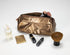 A metallic gold makeup bag with a zippered top, shown open with makeup brushes inside, alongside cosmetic items including a perfume bottle, compact powder, lipsticks, and a large makeup brush on a white background.