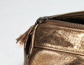 Close-up of a metallic bronze leather bag with a zipper and tassel detail on a light gray background.