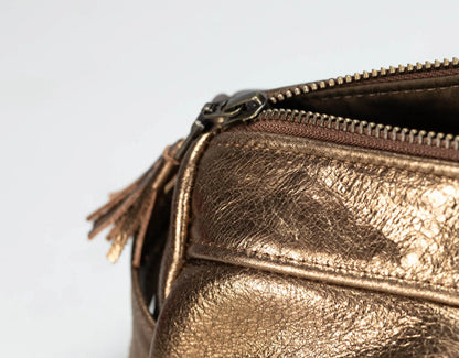 Close-up of a metallic bronze leather bag with a zipper and tassel detail on a light gray background.