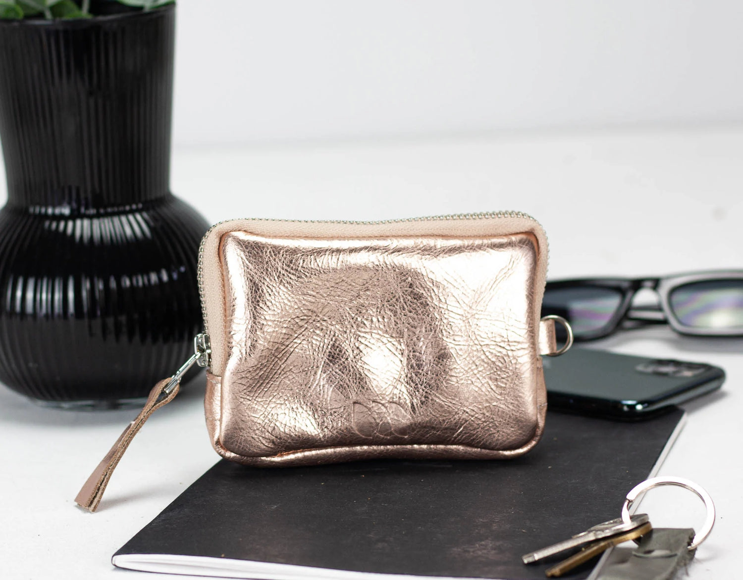 Rose gold pouch on a white surface with a plant and phone in the background