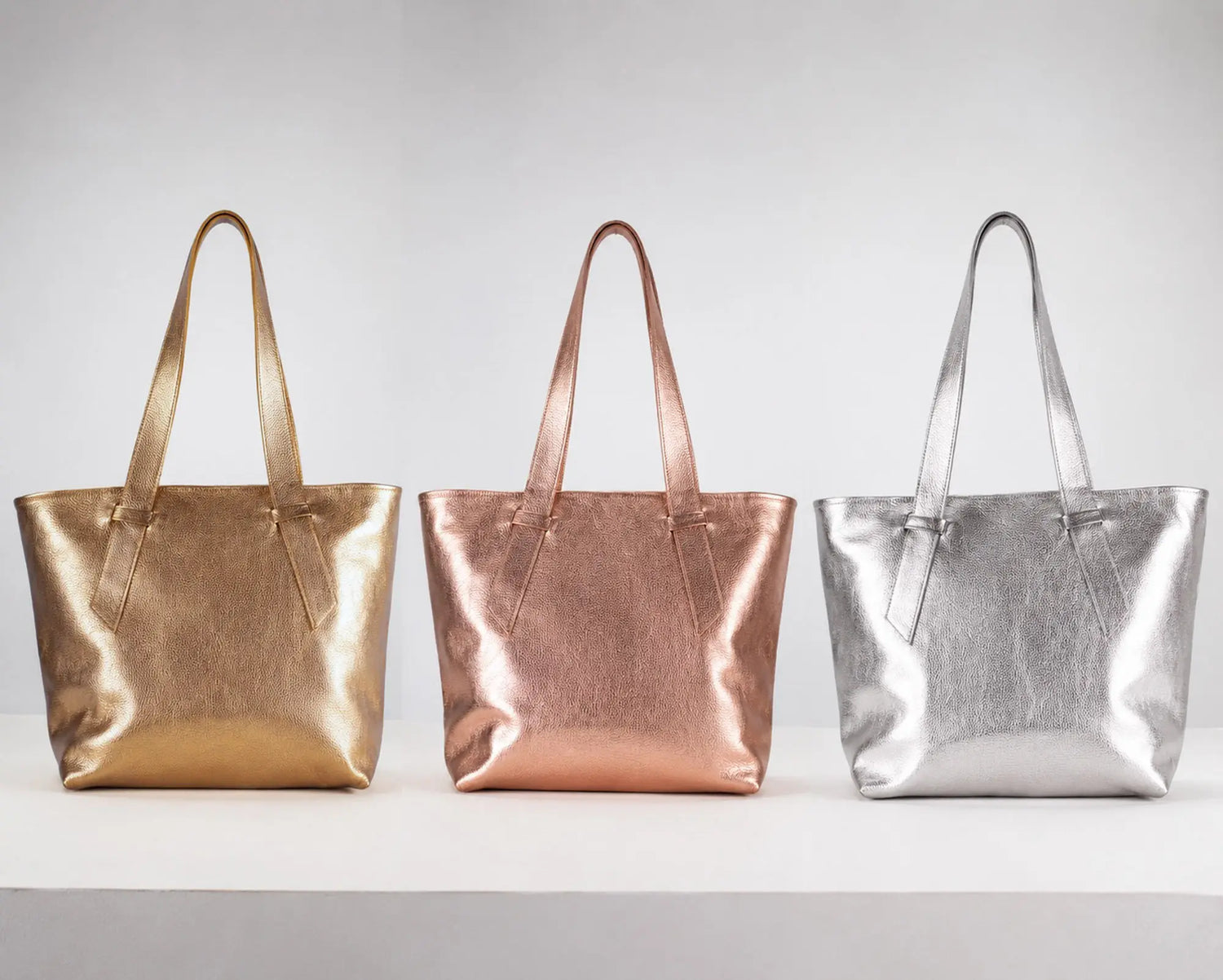 Three metallic tote bags in gold, rose gold, and silver, lined up side-by-side on a white surface against a light gray background.