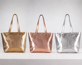 Three metallic tote bags in gold, rose gold, and silver, lined up side-by-side on a white surface against a light gray background.