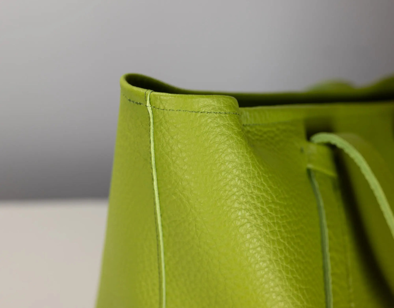 Close-up of a bright lime green pebbled leather bag corner with matching drawstring and visible stitching against a neutral gray background.