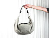 Gray handbag held by a hand against a white curtain background