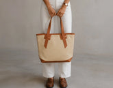 A person wearing white pants and brown leather loafers holds a large beige canvas tote bag with brown leather trim and handles against a neutral gray studio background.