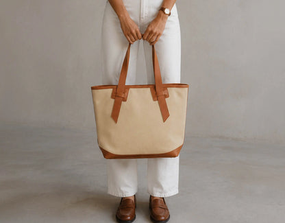 A person wearing white pants and brown leather loafers holds a large beige canvas tote bag with brown leather trim and handles against a neutral gray studio background.