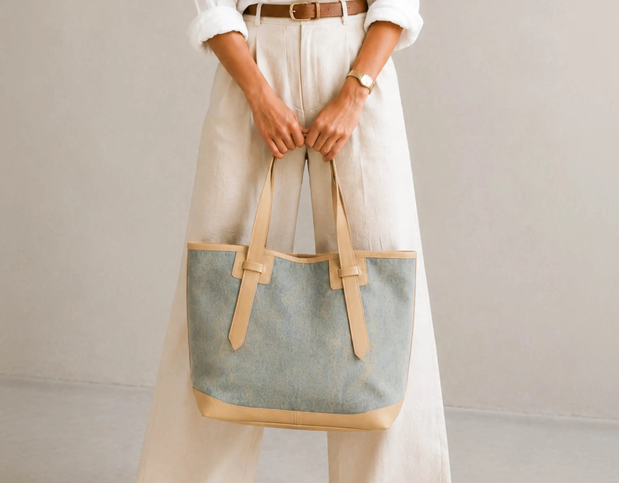 A person wearing cream wide-leg trousers and a white shirt holds a large canvas tote bag with a dusty blue body and tan leather trim and handles against a plain light gray background.