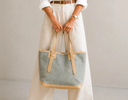 A person wearing cream wide-leg trousers and a white shirt holds a large canvas tote bag with a dusty blue body and tan leather trim and handles against a plain light gray background.