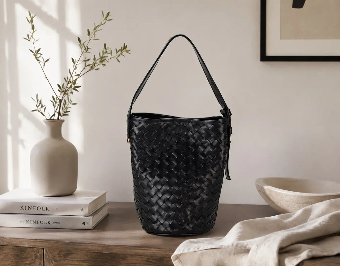 A black woven leather bucket bag with a single shoulder strap, displayed on a wooden table in a styled interior setting with a vase, books, and a linen cloth.