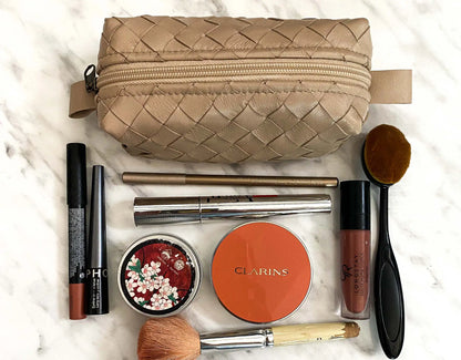 Makeup items including a Clarins product, lip gloss, and brushes on a marble surface with a woven beige pouch.