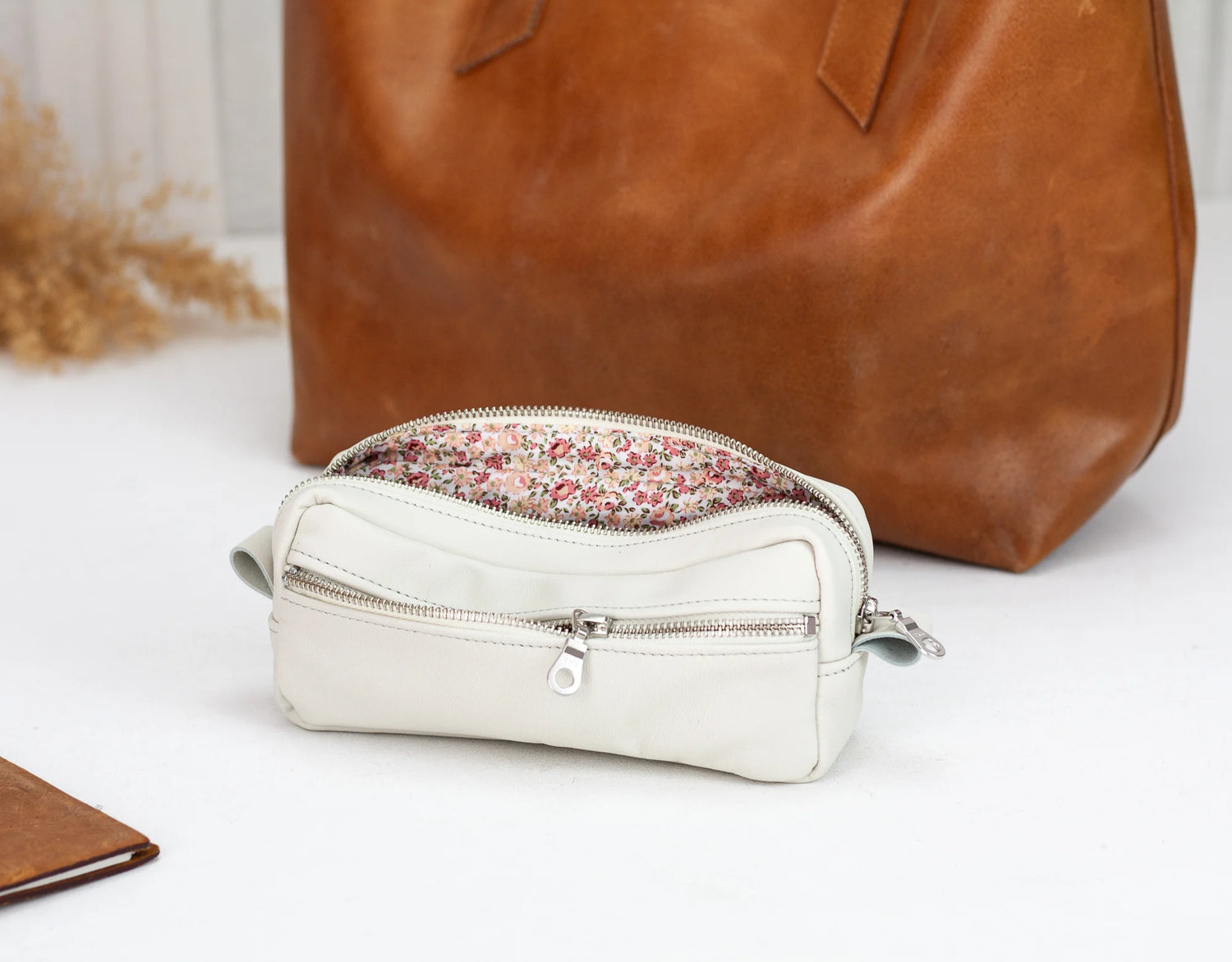 White leather pouch with floral zipper tab in front of a brown leather bag on a light background