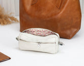 White leather pouch with floral zipper tab in front of a brown leather bag on a light background