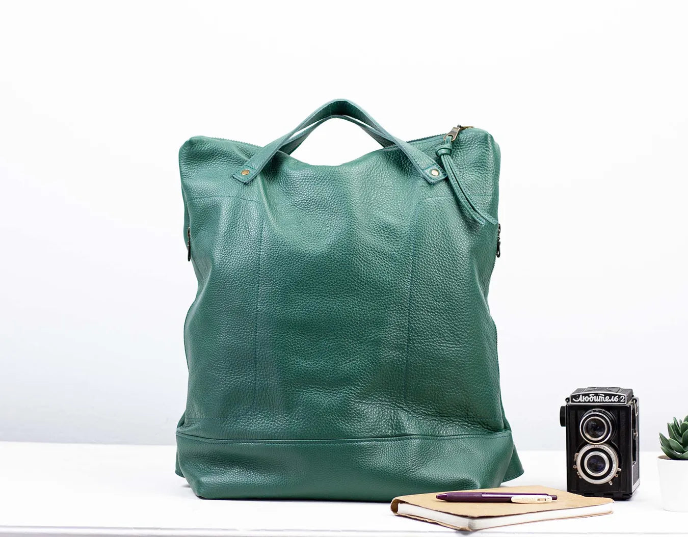 Green leather tote bag on a white background with a vintage camera and notebook.