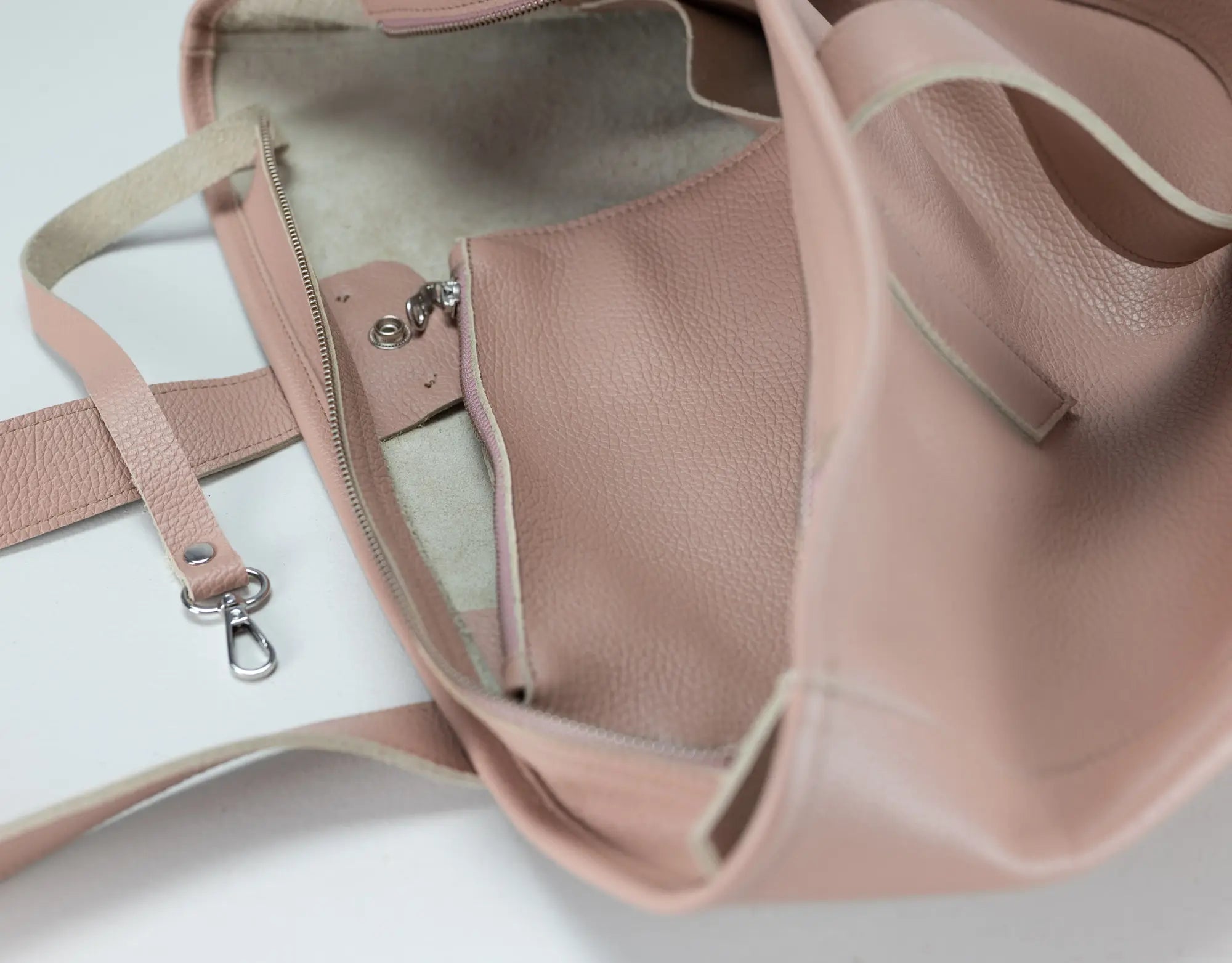 A close-up of a dusty pink pebbled leather tote bag shown open, displaying its interior lining, zipper pocket, and strap attachment on a white background.