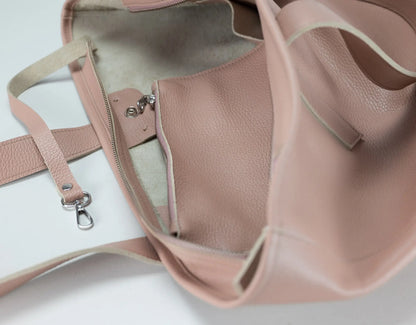 A close-up of a dusty pink pebbled leather tote bag shown open, displaying its interior lining, zipper pocket, and strap attachment on a white background.